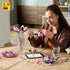 A woman assembles LEGO pink roses with glossy plastic petals and green stems, carefully shaping each curved bloom while seated at a wooden table in a bright home setting. Designed for detailed display with realistic plastic floral elements.