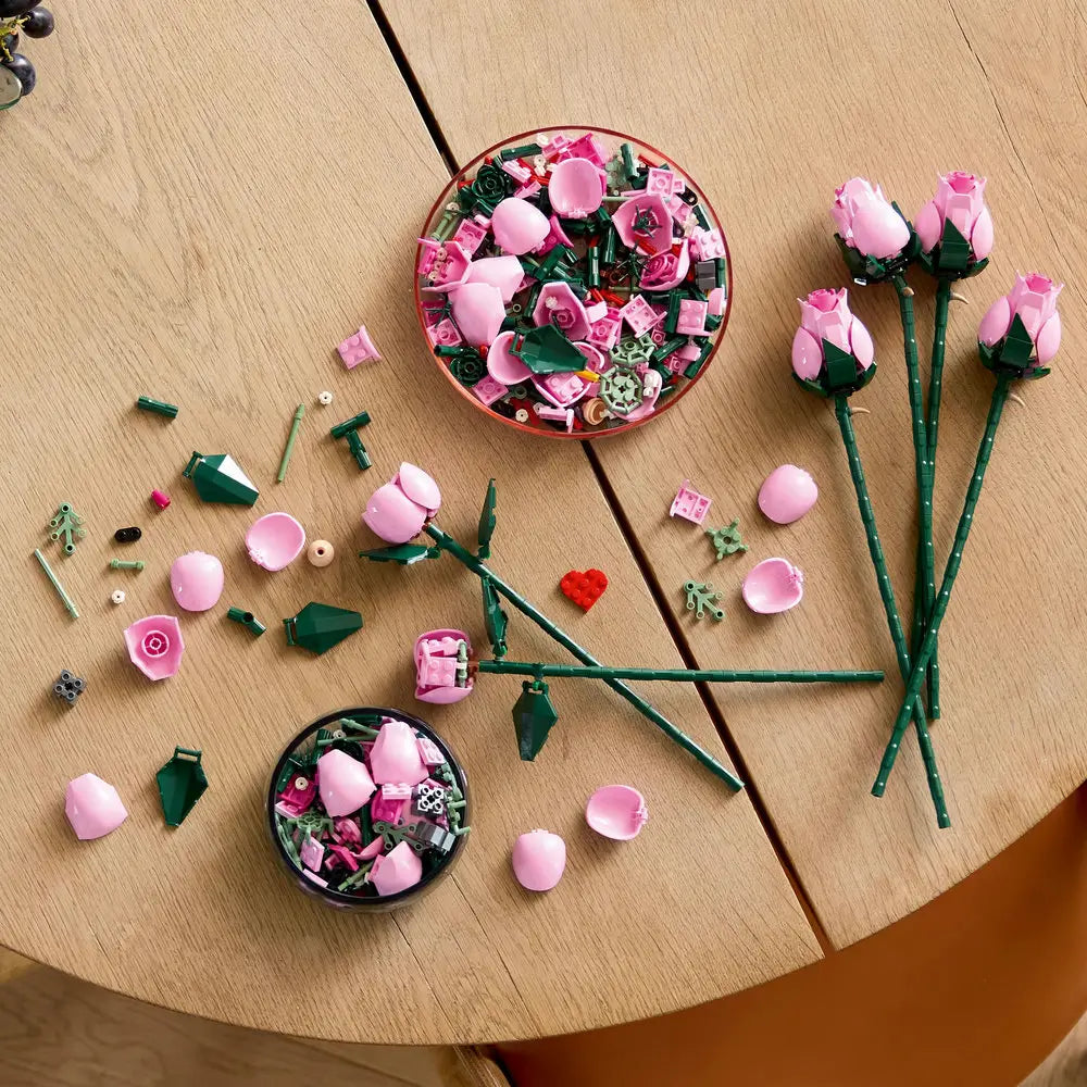 Loose LEGO pink rose pieces with smooth plastic petals and angular green stems are spread across a wooden table, showing the curved components arranged for creative floral assembly. Designed for detailed display with realistic plastic floral elements.