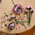 Loose LEGO pink rose pieces with smooth plastic petals and angular green stems are spread across a wooden table, showing the curved components arranged for creative floral assembly. Designed for detailed display with realistic plastic floral elements.