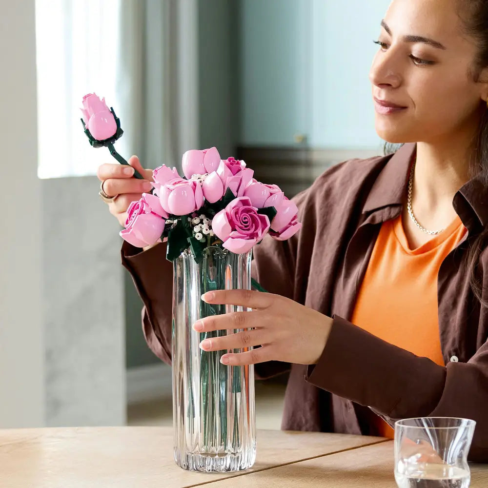 A woman arranges LEGO pink roses with curved plastic petals and long stems, placing the molded blooms into a tall clear glass vase on a wooden table in a softly lit room. Designed for detailed display with realistic plastic floral elements.