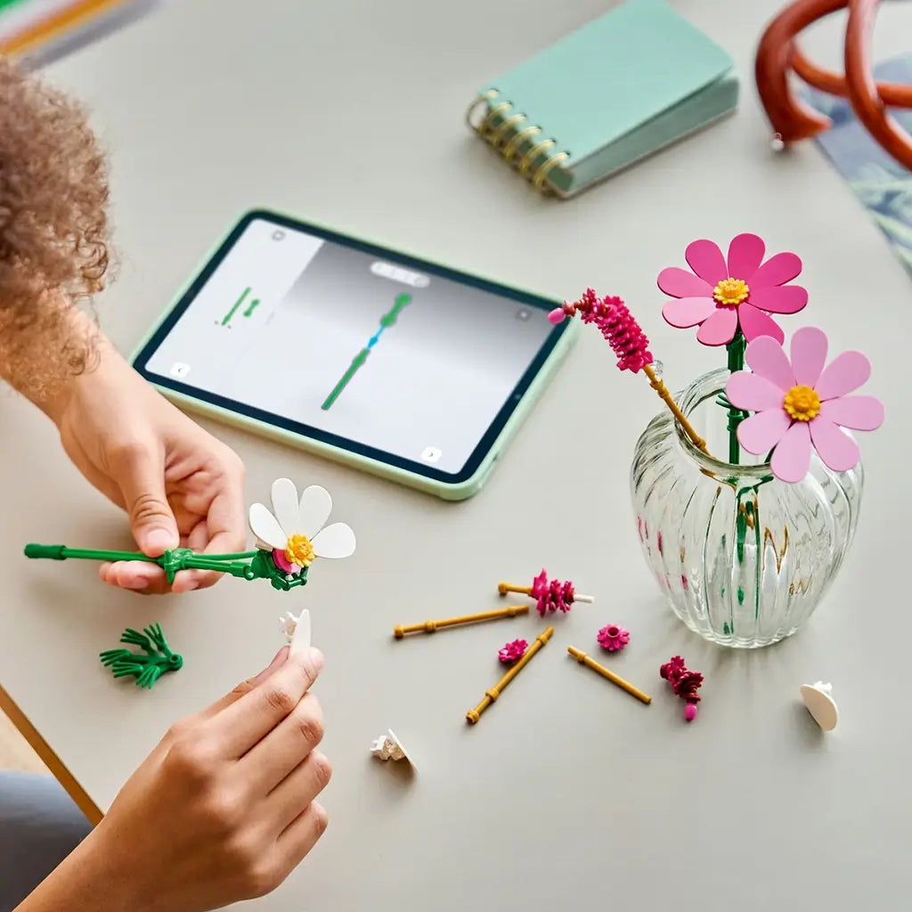 LEGO Botanicals Cosmos Flowers in pink, white, yellow, and green plastic are assembled by hand beside loose flower pieces, a clear vase, and a tablet on a tabletop.