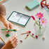 LEGO Botanicals Cosmos Flowers in pink, white, yellow, and green plastic are assembled by hand beside loose flower pieces, a clear vase, and a tablet on a tabletop.