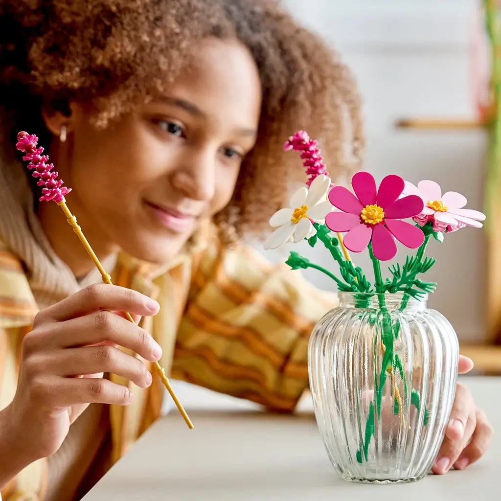 LEGO Botanicals Cosmos Flowers in pink, magenta, white, yellow, and green plastic fill a clear glass vase as a person adds a tall flower stem at a white tabletop.