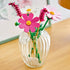 LEGO Botanicals Cosmos Flowers in pink, magenta, white, yellow, and green plastic are arranged as a decorative bouquet in a ribbed clear glass vase on a wood table.