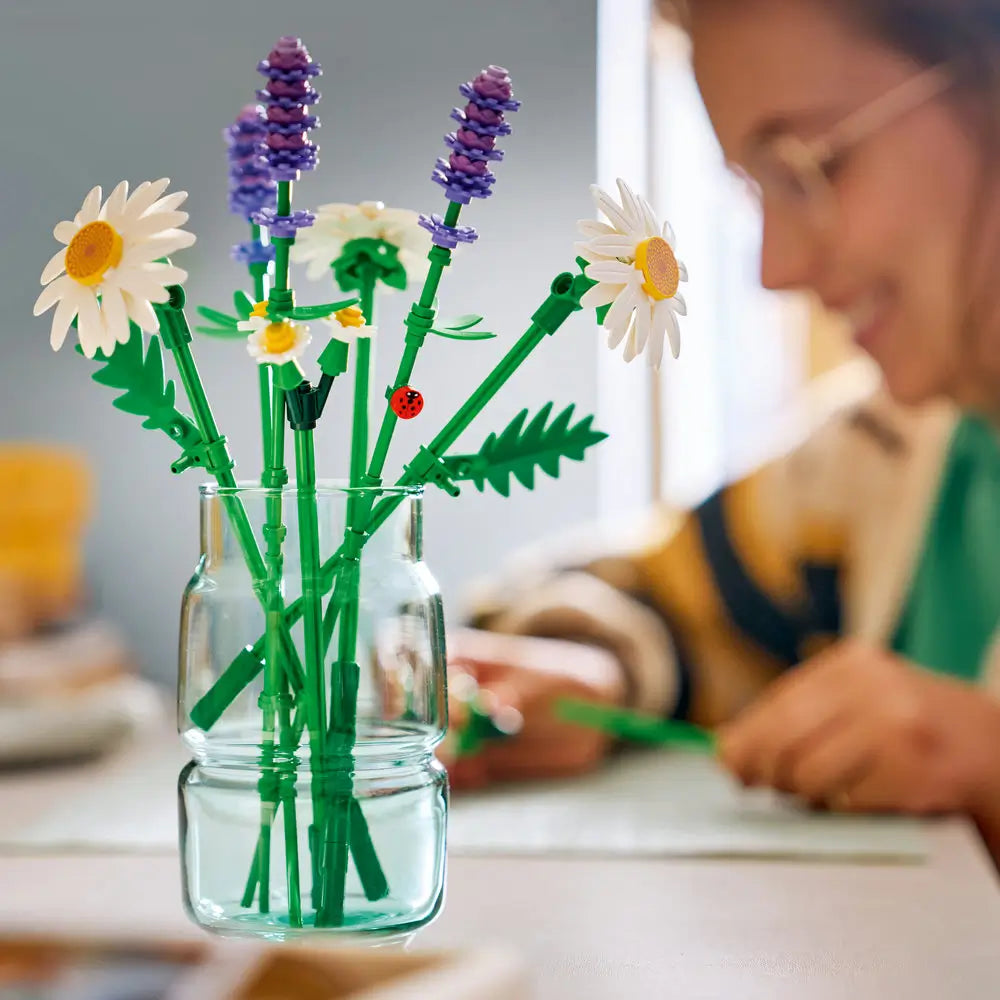 LEGO Botanicals Daisies lifestyle image showing white and yellow plastic daisy flowers with purple lavender accents, slender green stems for home decor, displayed in a clear glass jar on a tabletop.