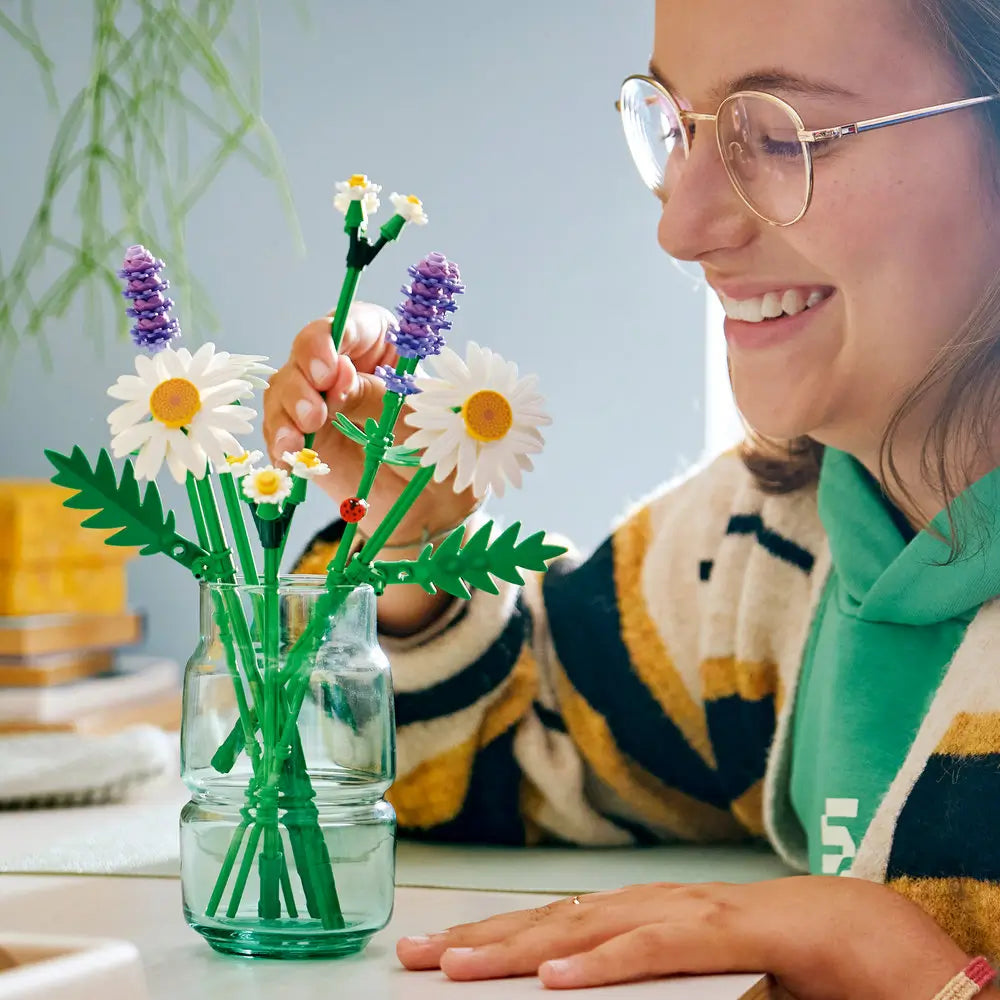 LEGO Botanicals Daisies lifestyle builder image showing hands assembling green plastic stems and white daisy petals, fine botanical shapes for decorative display, with small flower elements arranged on a mat.