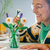 LEGO Botanicals Daisies lifestyle builder image showing hands assembling green plastic stems and white daisy petals, fine botanical shapes for decorative display, with small flower elements arranged on a mat.
