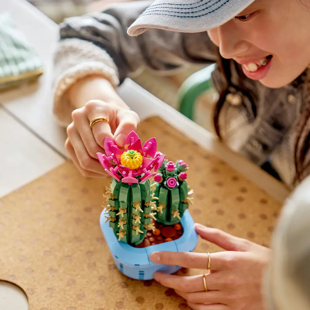 LEGO Botanicals Flowering Cactus lifestyle image featuring green textured plastic cactus stems with beige spines and pink blossoms, rounded sculptural form for home decor, shown being gently adjusted on a tabletop.