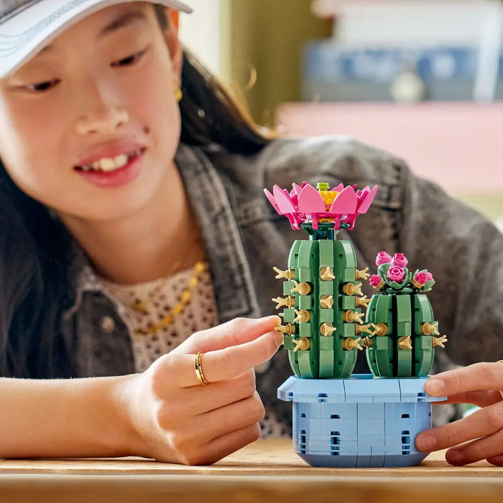 LEGO Botanicals Flowering Cactus lifestyle builder image showing hands assembling green plastic cactus sections, textured cylindrical shapes for decorative display, surrounded by pink flower elements and small brick pieces.