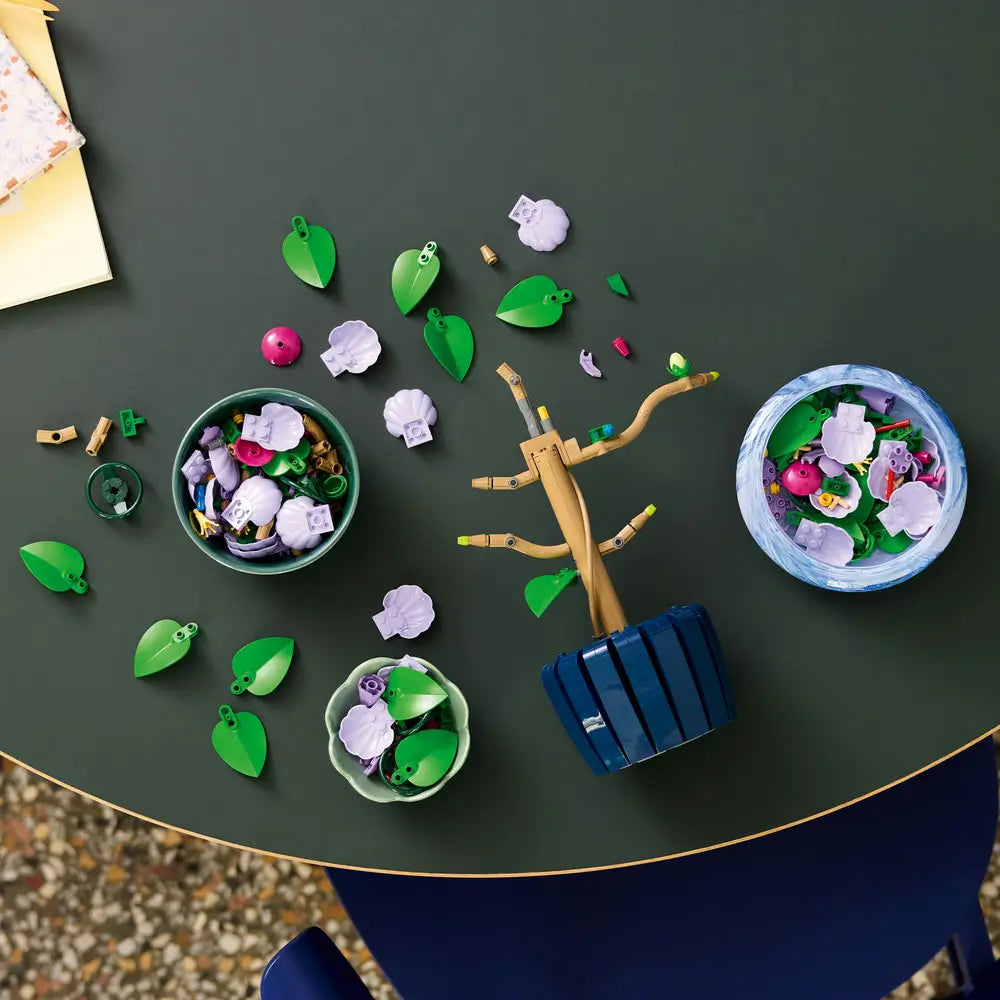 Overhead view of LEGO Botanicals Hibiscus build in progress, with tan branches, green leaf pieces, and purple petals spread across bowls on a dark tabletop.