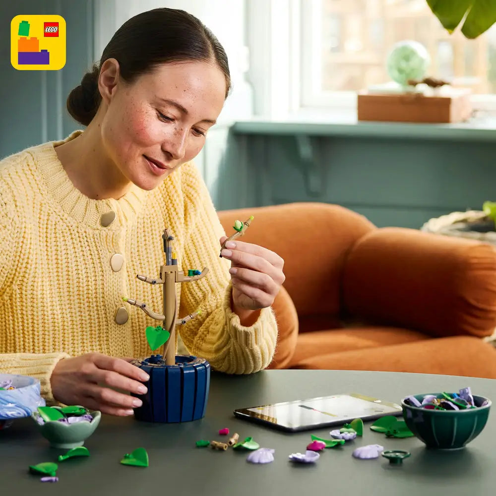 Person assembling the LEGO Botanicals Hibiscus set, carefully attaching green leaves to a tan stem in a blue pot, surrounded by colorful LEGO flower pieces and digital instructions.
