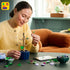 Person assembling the LEGO Botanicals Hibiscus set, carefully attaching green leaves to a tan stem in a blue pot, surrounded by colorful LEGO flower pieces and digital instructions.