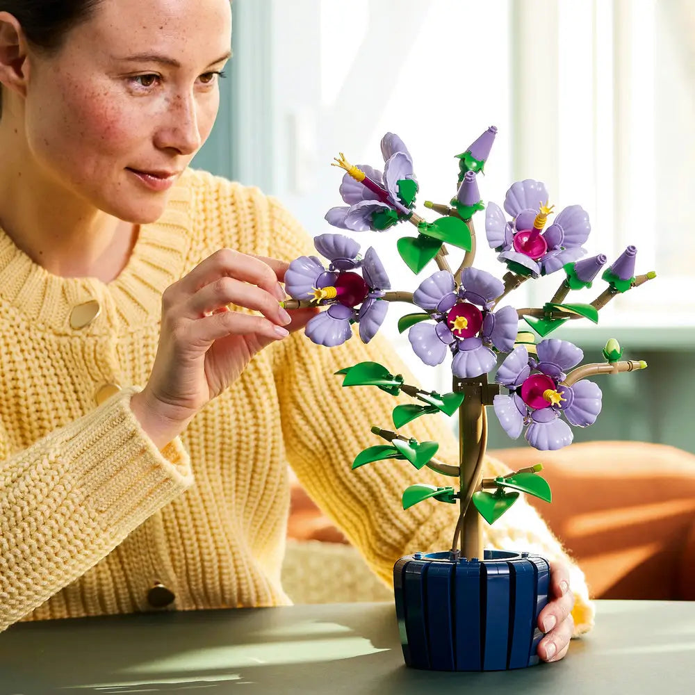 Builder in a yellow sweater adding small LEGO leaf pieces to the Botanicals Hibiscus plant, with parts and instructions neatly arranged on the table.