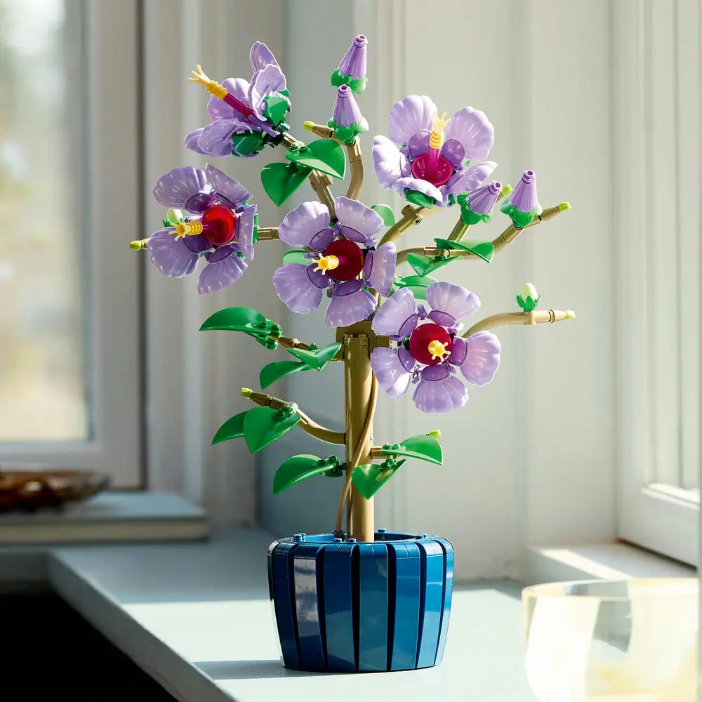 Completed LEGO Botanicals Hibiscus model with purple and magenta blossoms, green leaves, and a golden stem displayed on a windowsill in natural light.