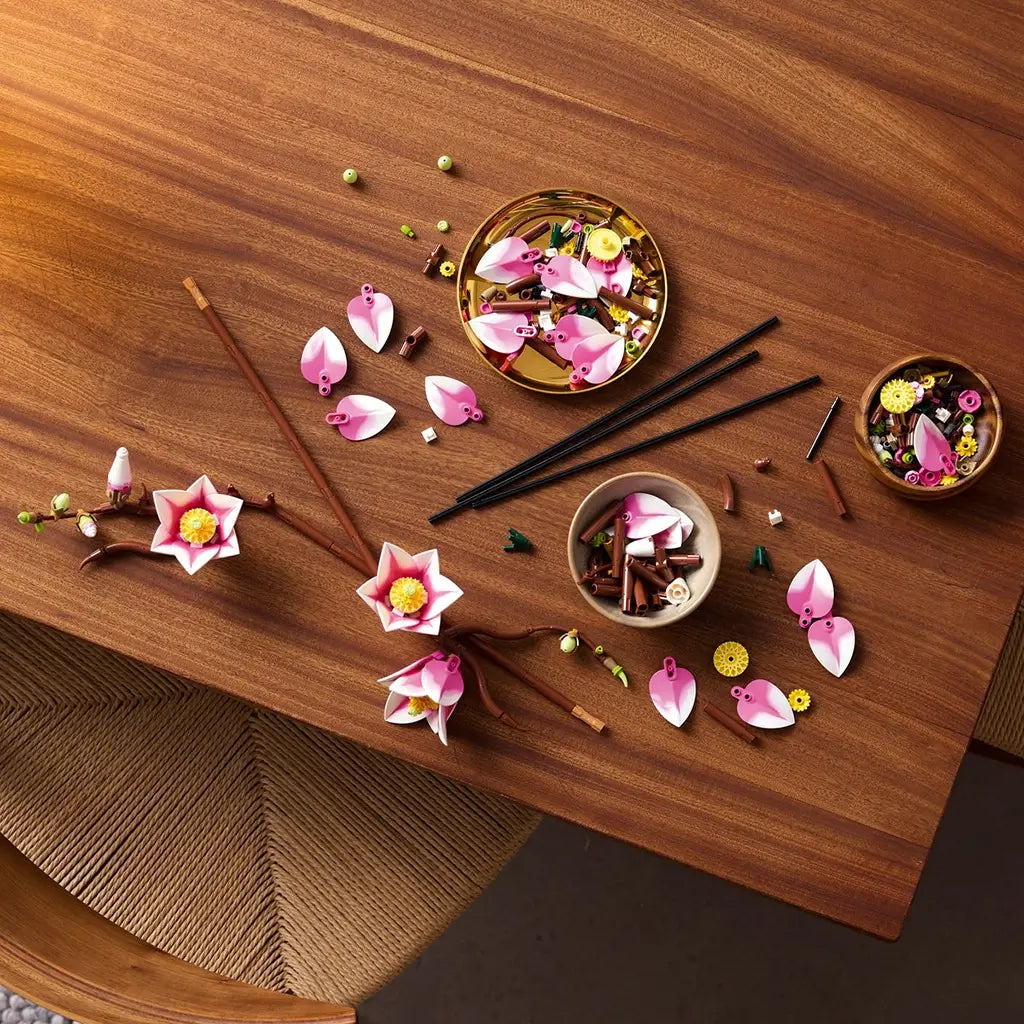 LEGO magnolia branches build features pink and white plastic petals, brown stems, and small green buds, arranged as a floral assembly project on a wooden table with tools and parts in a home crafting setting.