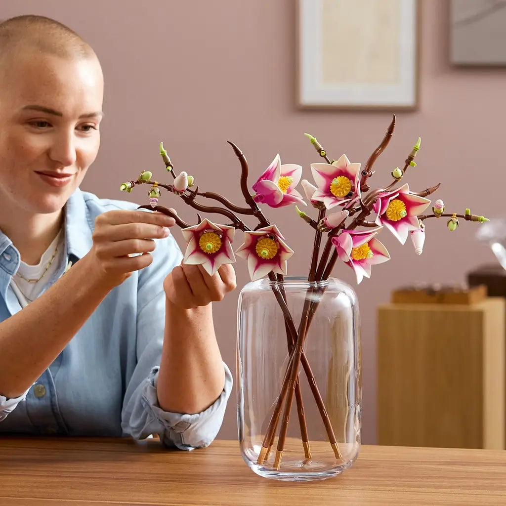 LEGO magnolia branches model in use shows a person arranging pink and white plastic blossoms on brown stems in a clear glass vase, demonstrating decorative home styling in a warm indoor setting.