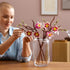 LEGO magnolia branches model in use shows a person arranging pink and white plastic blossoms on brown stems in a clear glass vase, demonstrating decorative home styling in a warm indoor setting.