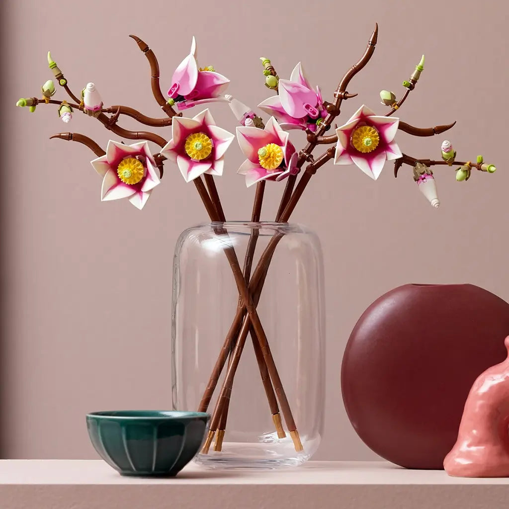 LEGO magnolia branch display features pink and white plastic flowers with yellow centers on brown stems in a clear glass vase, styled as modern floral home decor on a tabletop with minimal background.