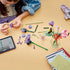 LEGO Botanicals Olivia Rodrigo's Flower Bouquet pieces in purple, pink, red, and green plastic cover a tan table as a builder assembles blooms beside a tablet.