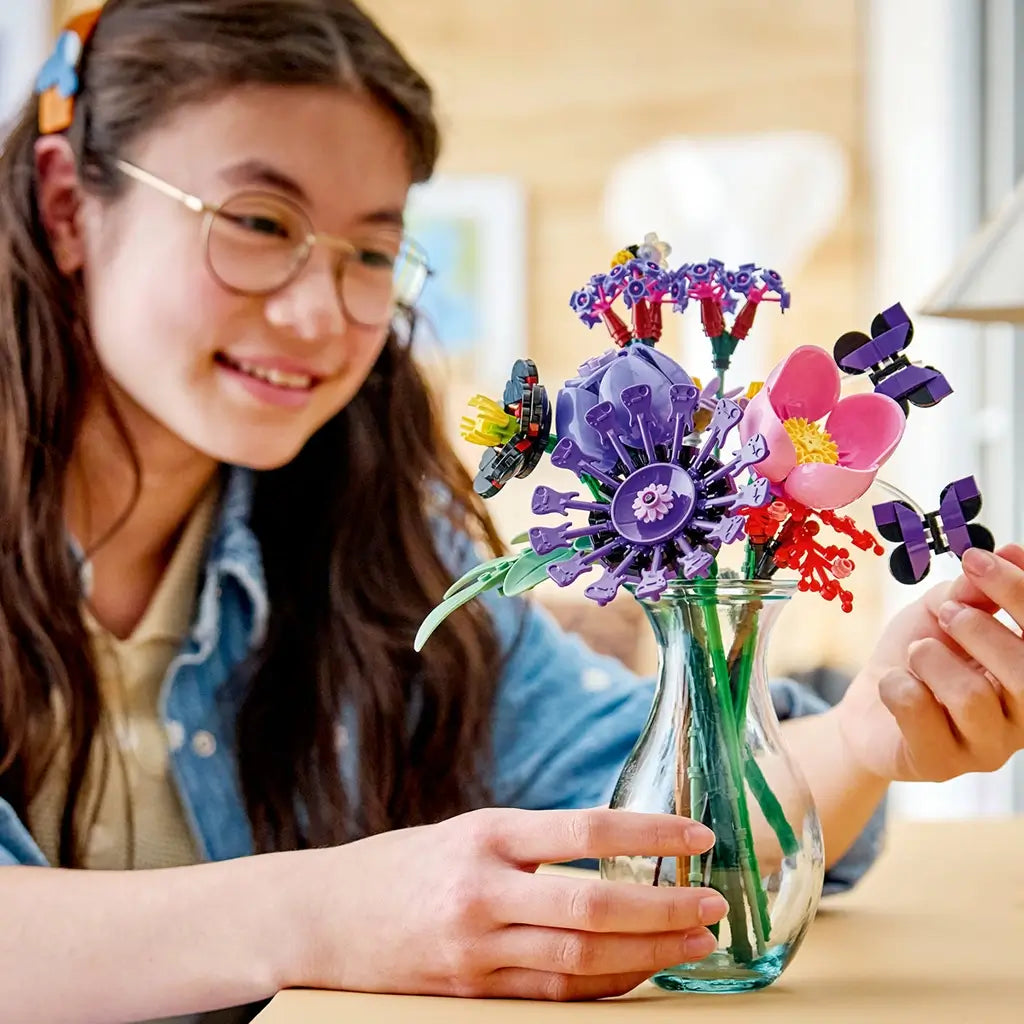 LEGO Botanicals Olivia Rodrigo's Flower Bouquet in purple, pink, red, green, and black plastic fills a clear vase as a smiling builder holds a butterfly stem.