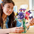LEGO Botanicals Olivia Rodrigo's Flower Bouquet in purple, pink, red, green, and black plastic fills a clear vase as a smiling builder holds a butterfly stem.