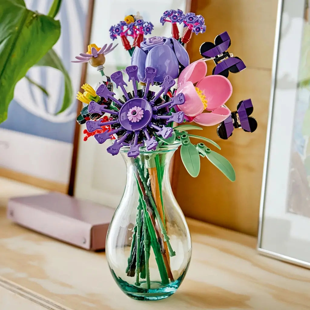 LEGO Botanicals Olivia Rodrigo's Flower Bouquet features purple, pink, red, green, and black plastic blooms with butterflies arranged in a clear glass vase by a window.