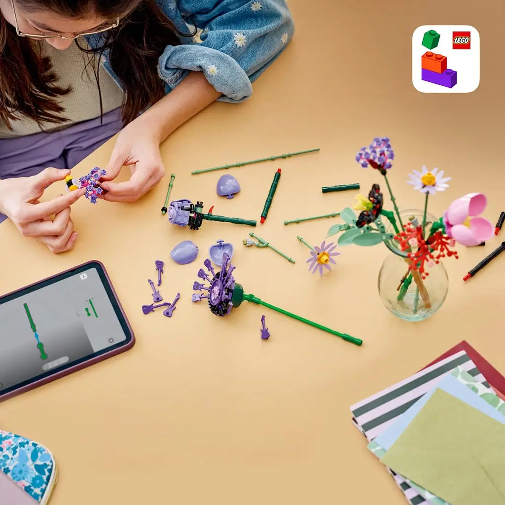 LEGO Botanicals Olivia Rodrigo's Flower Bouquet pieces in purple, pink, red, and green plastic are assembled on a tan table beside a tablet and a partly filled vase.