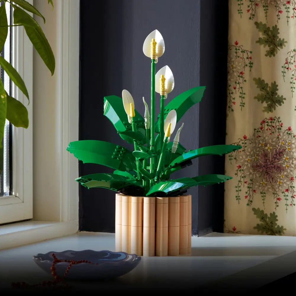 LEGO Botanicals Peace Lily lifestyle image showing deep green plastic leaves and white flower spathes, smooth curved botanical forms for home decor, placed in a tan brick planter on a windowsill with natural light.