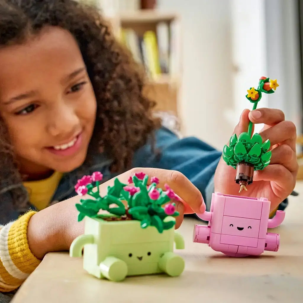 LEGO Botanicals Rocking Plants set in pink and light green plastic bricks shows a child lifting leafy flower inserts from smiling planters on a light wood table.
