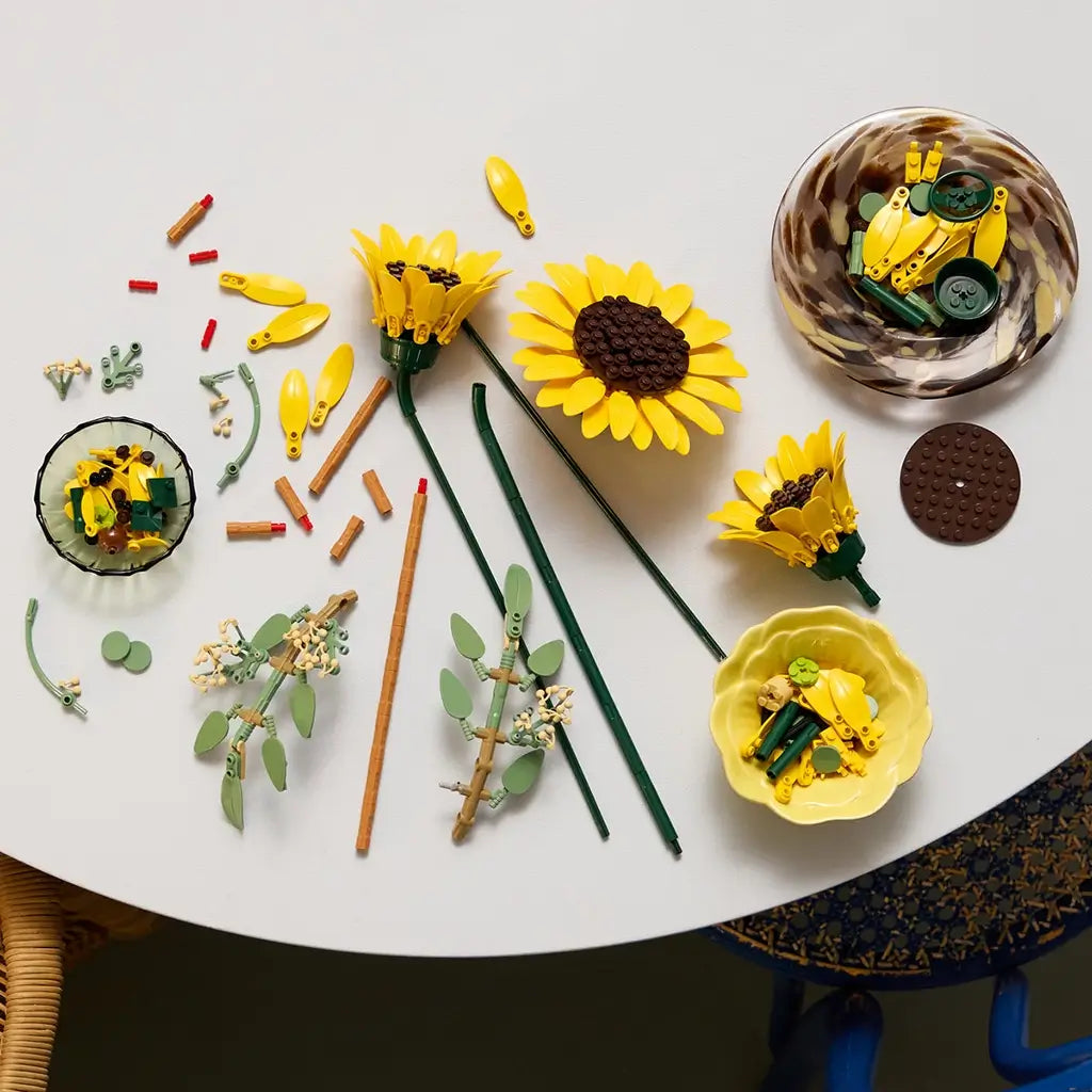 LEGO sunflower bouquet build process shows yellow plastic petals, green stems, and small connector pieces spread across a table, illustrating step-by-step assembly of decorative floral stems in a crafting workspace.