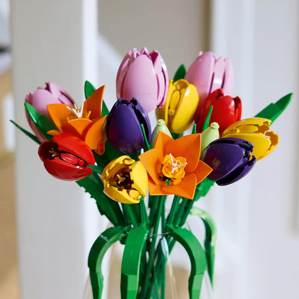 LEGO tulip bouquet arranged in a clear glass vase shows multicolored plastic flowers with glossy curved petals and green stems, displayed upright as home décor against a softly lit indoor background.