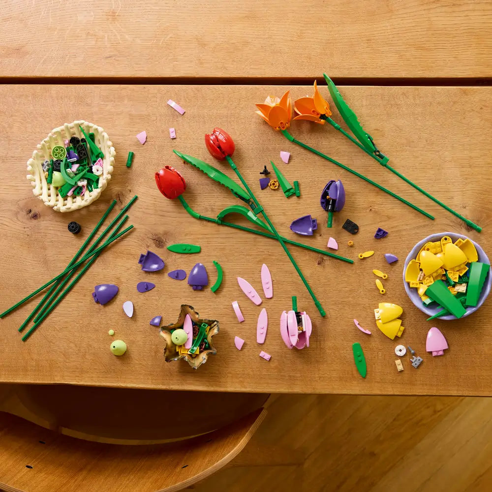 LEGO tulip bouquet building process shows assorted colorful plastic petals, green stems, and connectors spread across a wooden table, illustrating the construction stages of the decorative floral set.
