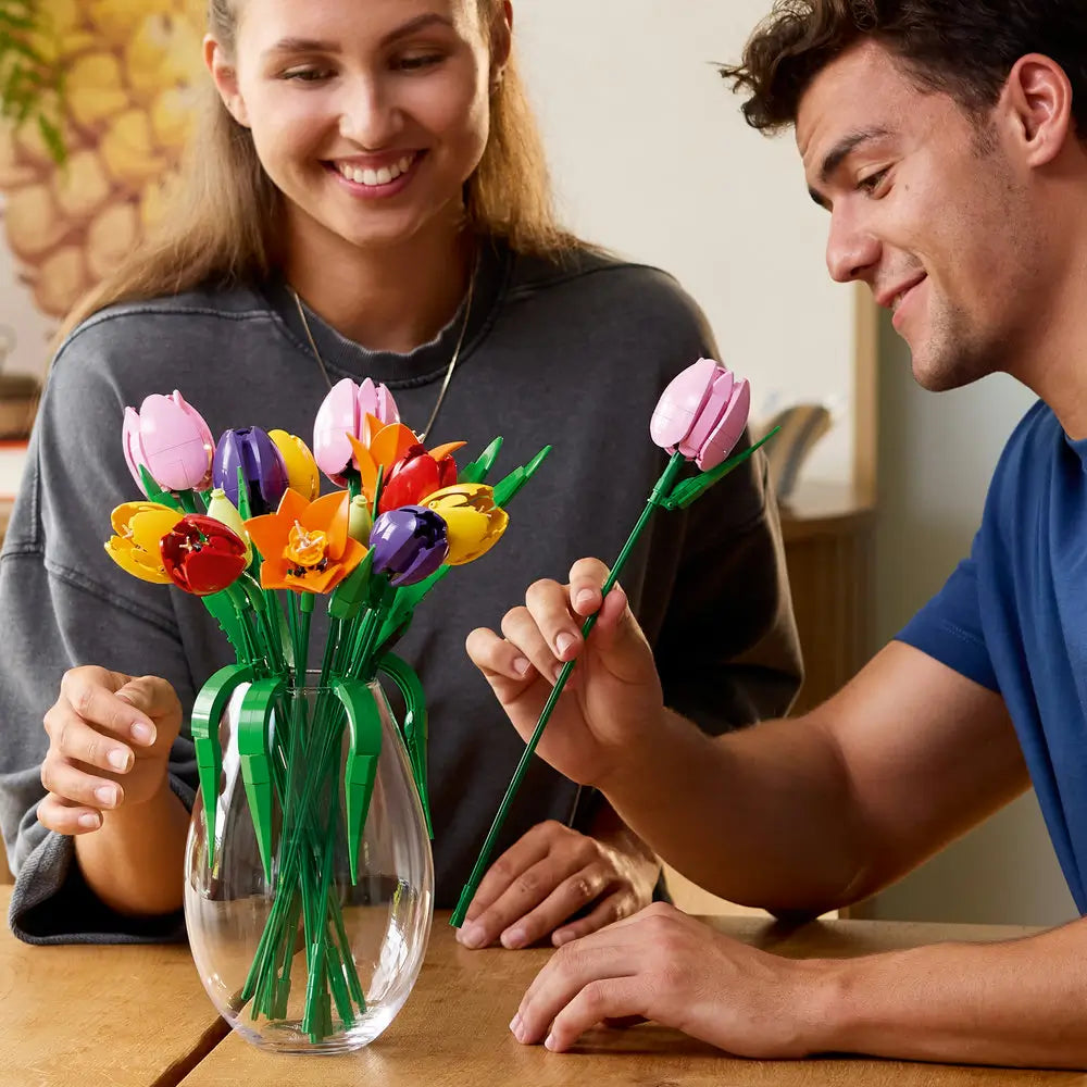 LEGO tulip bouquet lifestyle scene shows two adults smiling while arranging colorful plastic tulip stems in a clear glass vase, demonstrating the interactive building and display experience at a home table.
