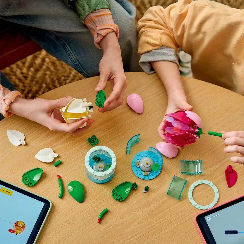 LEGO Botanicals Water Lilies in pink, white, green, and clear plastic are assembled by children from loose petals, stems, and round base pieces on a wood table.