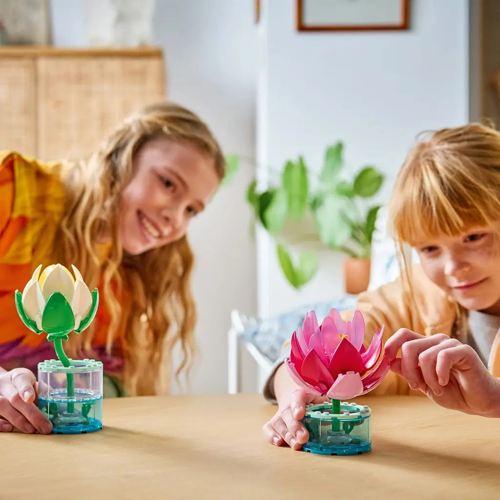 LEGO Botanicals Water Lilies in white, yellow, pink, purple, green, and clear plastic sit on a wood table as two children adjust the layered flower models indoors.