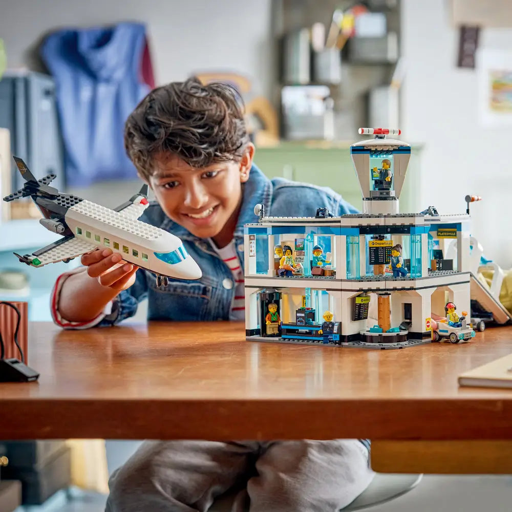 LEGO City airport terminal and airplane playset held by a child, featuring blue structural supports, clear control tower, and passenger jet, designed for interactive roleplay and storytelling.