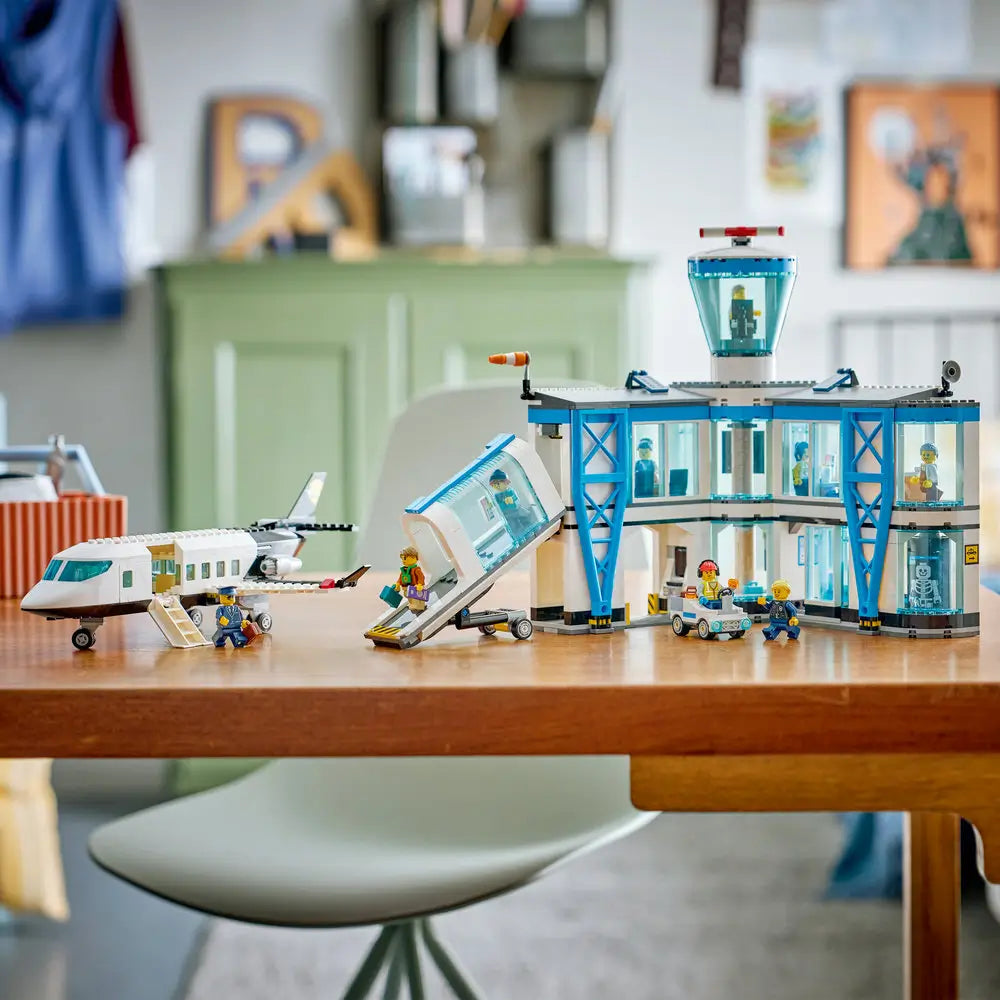LEGO City airport playset fully assembled on a table, showing plastic terminal building, jet bridge, airplane, and minifigures arranged to simulate airport operations and travel scenes.
