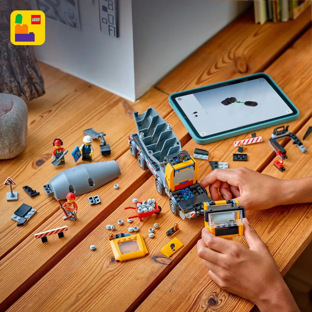 LEGO City cement mixer build scene showing yellow and gray plastic truck parts, loose bricks, and minifigures spread on a wooden table, with a tablet displaying digital instructions nearby.