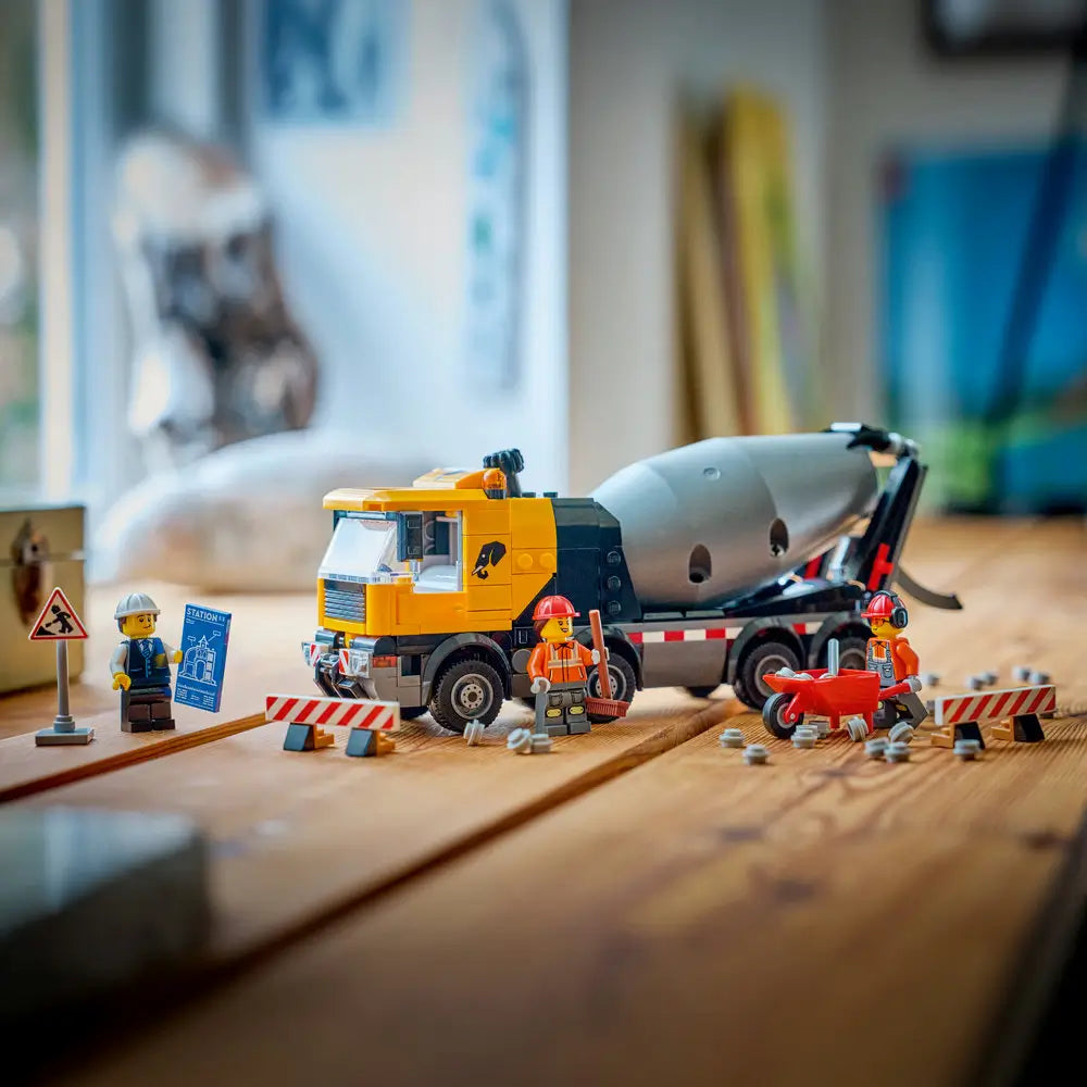 LEGO City cement mixer toy scene with yellow plastic truck, gray rotating drum, minifig workers, and striped barriers arranged on a tabletop to represent an active construction site setting.