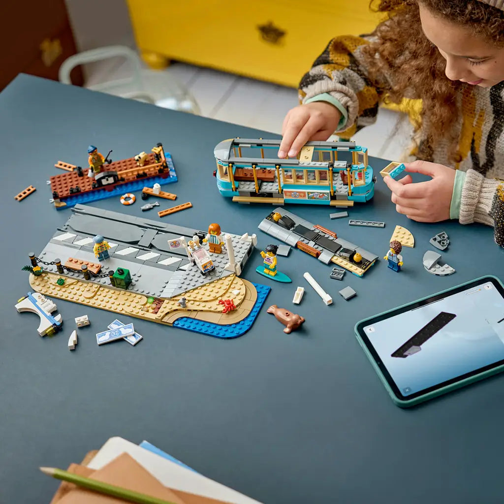 LEGO City Classic Beach Streetcar in turquoise, tan, gray, and brown plastic bricks is assembled by a child with a seaside platform, pier, minifigures, and tablet.