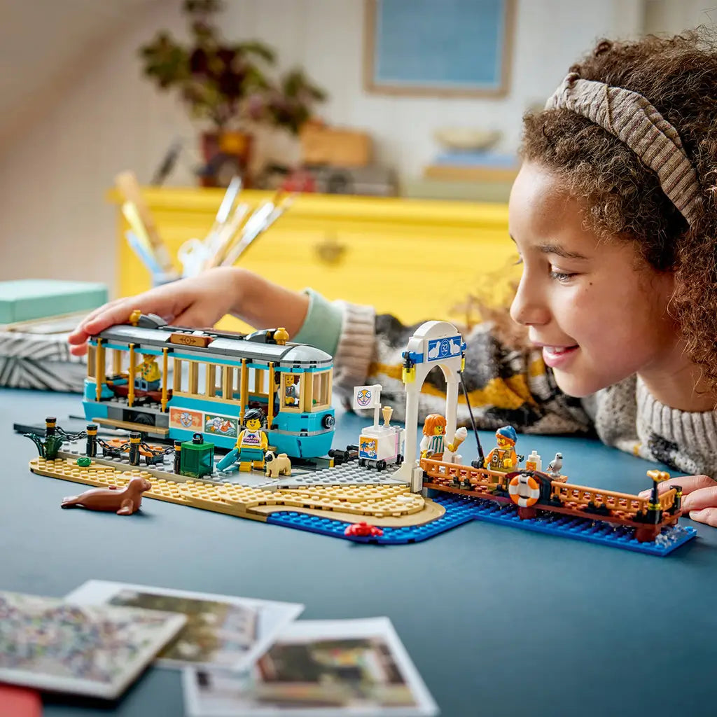 LEGO City Classic Beach Streetcar in turquoise, tan, blue, and brown plastic bricks sits by a beach platform and fishing pier as a child plays with the seaside scene.