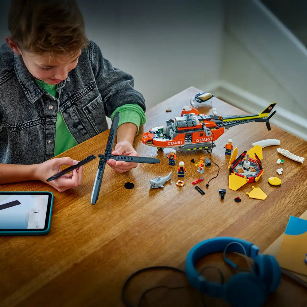 LEGO City building activity scene showing a child assembling the Coast Guard helicopter on a wooden table, with rotor blades, lifeboat pieces, minifigures, and digital instructions visible on a tablet nearby.