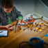 LEGO City building activity scene showing a child assembling the Coast Guard helicopter on a wooden table, with rotor blades, lifeboat pieces, minifigures, and digital instructions visible on a tablet nearby.