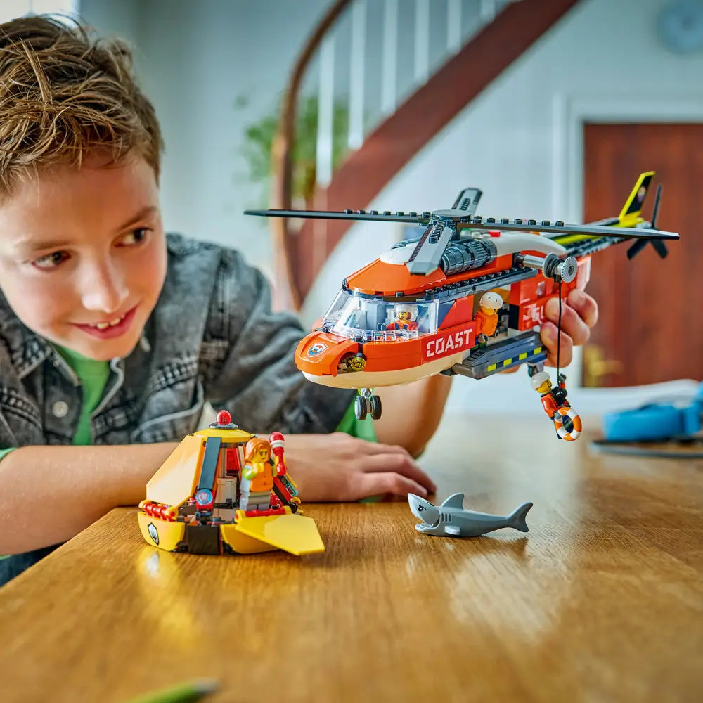 LEGO City lifestyle image of a child playing with the Coast Guard helicopter rescue set, lifting the aircraft and lifeboat pod while acting out ocean rescue scenarios in a bright indoor home setting.