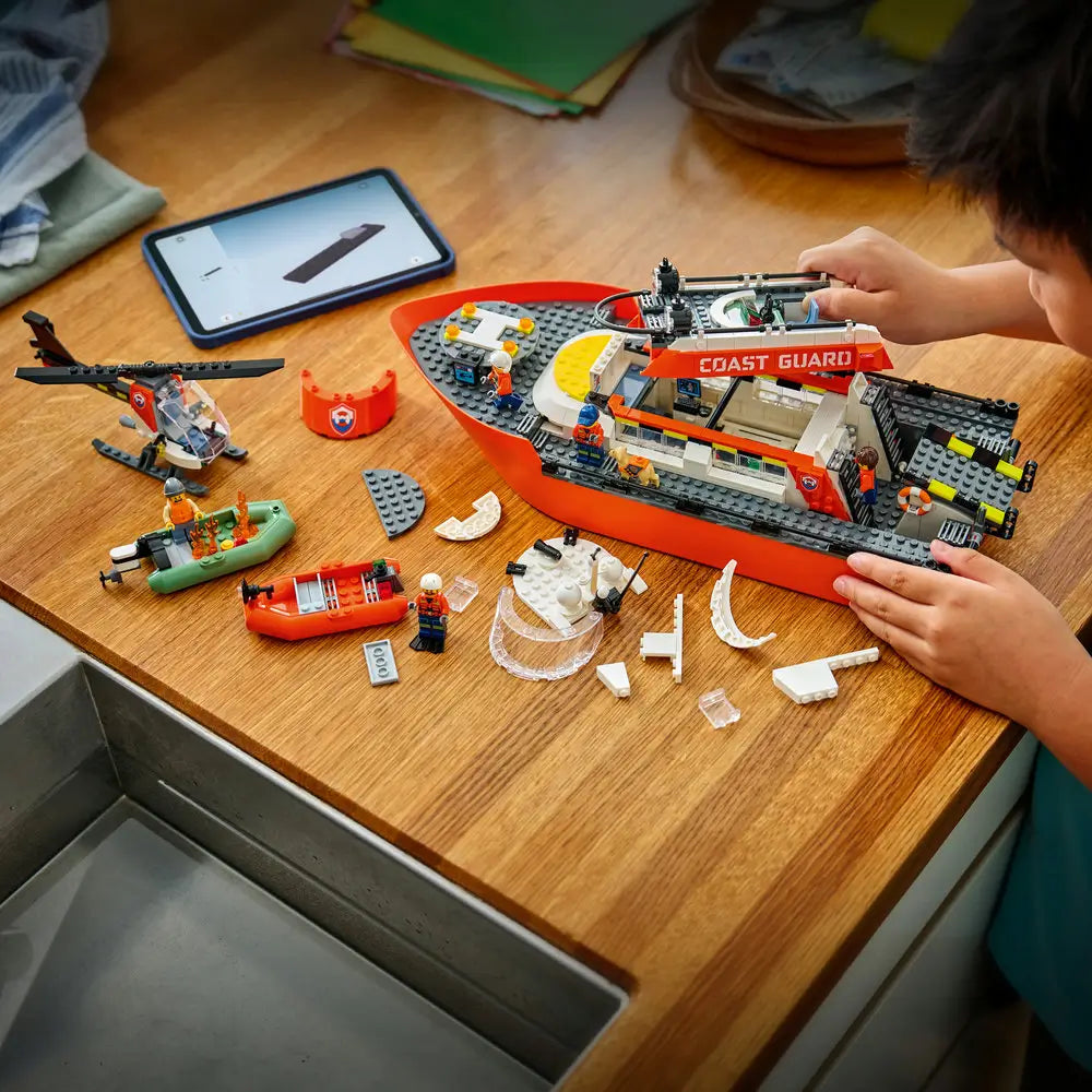 LEGO City building activity scene showing a child assembling the Coast Guard rescue boat on a wooden table, with scattered bricks, helicopter parts, minifigures, and digital instructions visible on a tablet.