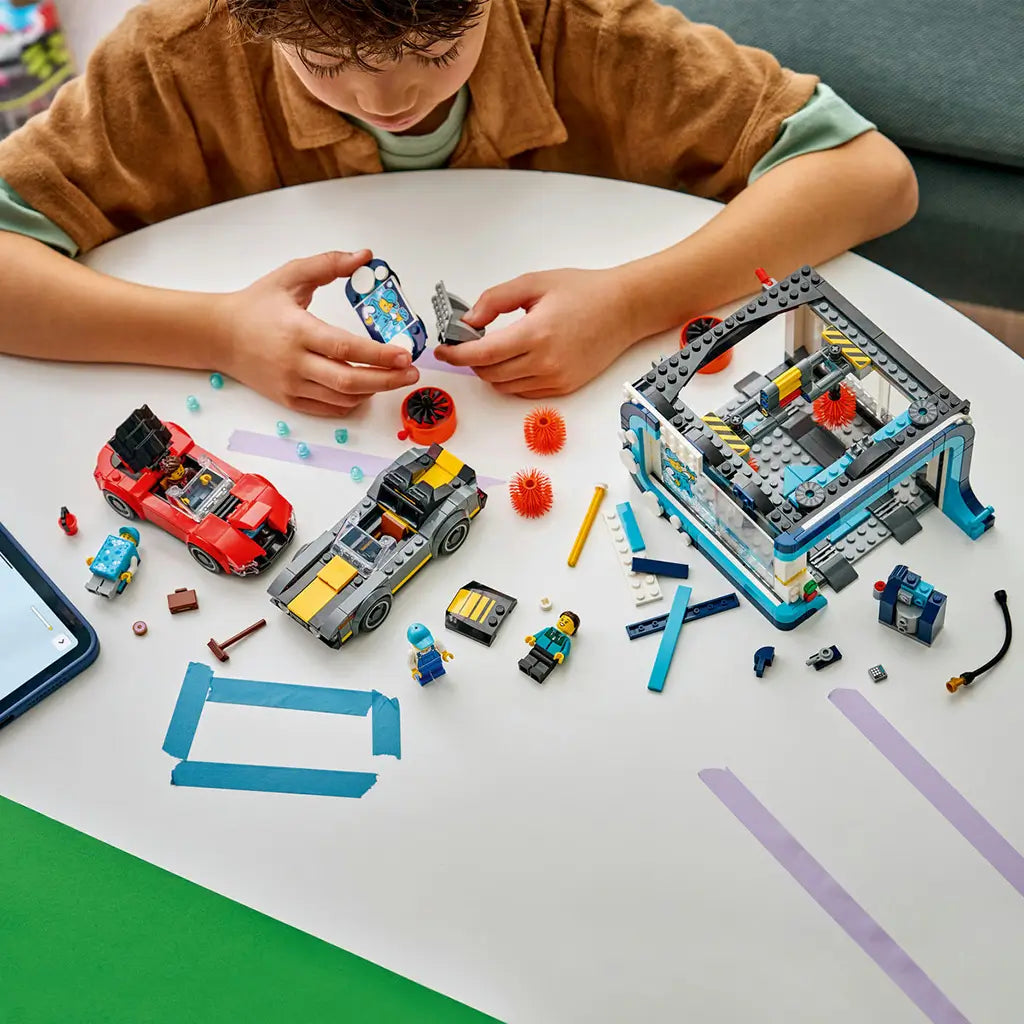 LEGO City Drive-Through Car Wash in teal, white, red, and gray plastic bricks is assembled by a child at a table with two cars, minifigures, brushes, and loose pieces.