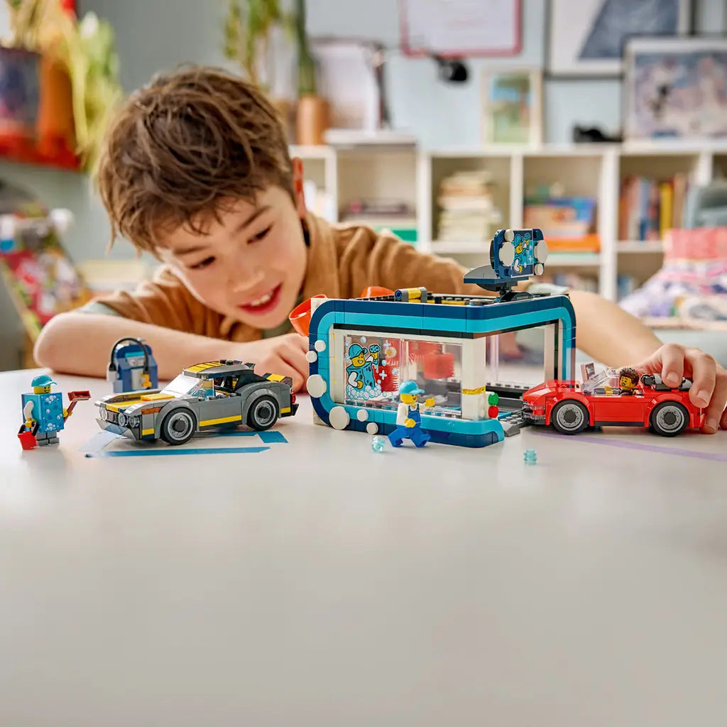 LEGO City Drive-Through Car Wash in teal, white, red, gray, and yellow plastic bricks is played with by a child, with two cars, wash brushes, bubbles, and minifigures.