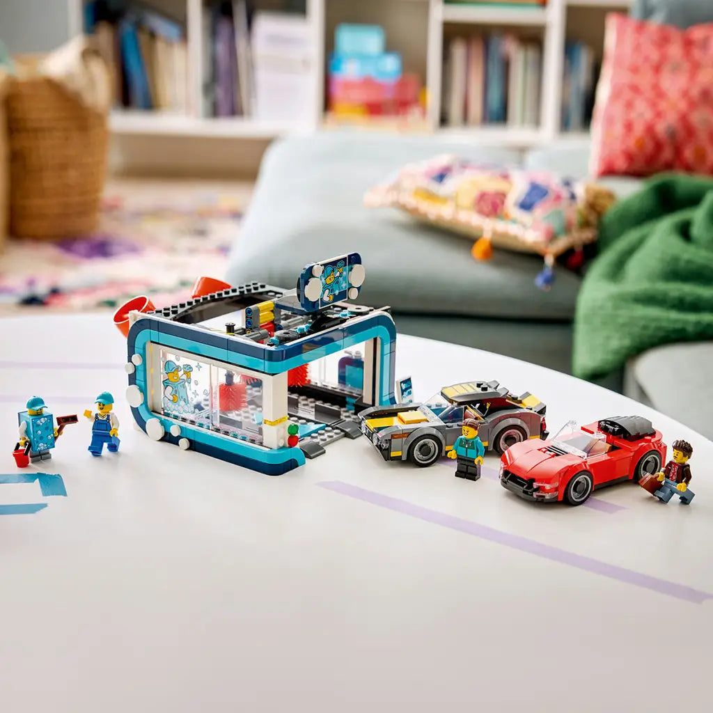 LEGO City Drive-Through Car Wash in teal, white, red, gray, and yellow plastic bricks is displayed with two cars and four minifigures on a table in a living room.