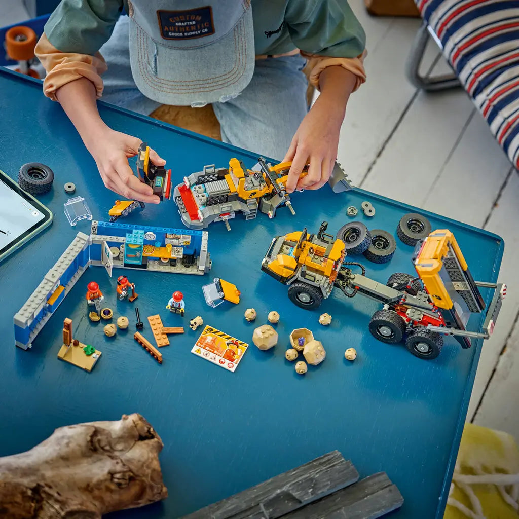 LEGO City Dump Truck & Front End Loader in yellow, black, gray, and blue plastic bricks is assembled by a child on a blue table with loose wheels, rocks, workers, and site office.