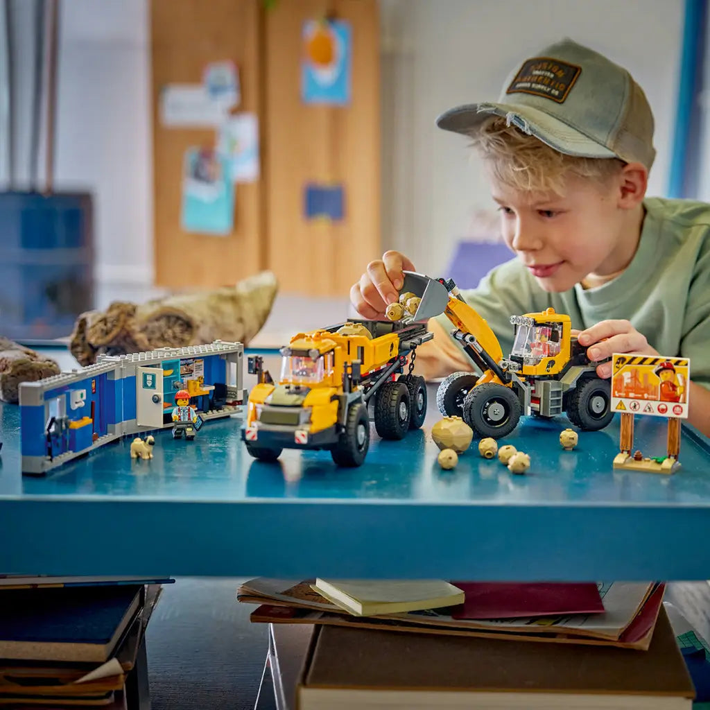 LEGO City Dump Truck & Front End Loader in yellow and black plastic bricks is played with by a child, who loads tan rocks beside a blue site office, workers, safety sign, and dog.